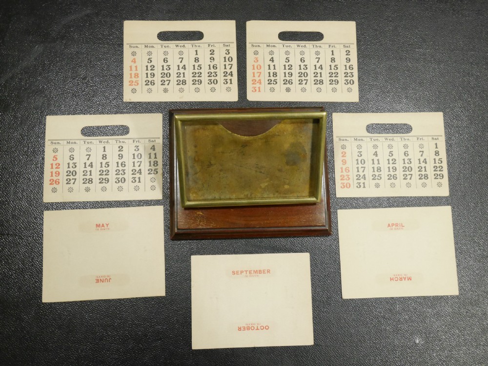 A vintage perpetual desk calendar with a polished brass frame holding paper month cards, mounted on a dark mahogany wooden base. View VII