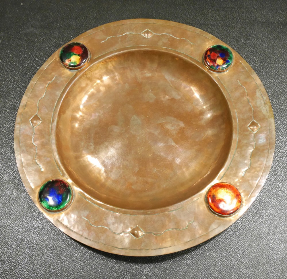 A circular hand-hammered copper charger with a sunken central dish, chased border decoration, and four inset colourful enamel cabochons.