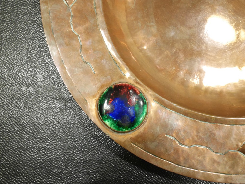 A circular hand-hammered copper charger with a sunken central dish, chased border decoration, and four inset colourful enamel cabochons. View II