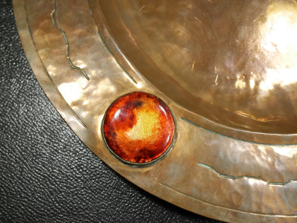 A circular hand-hammered copper charger with a sunken central dish, chased border decoration, and four inset colourful enamel cabochons. View III