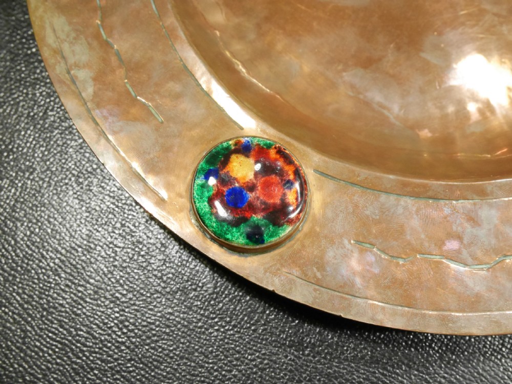 A circular hand-hammered copper charger with a sunken central dish, chased border decoration, and four inset colourful enamel cabochons. View IV