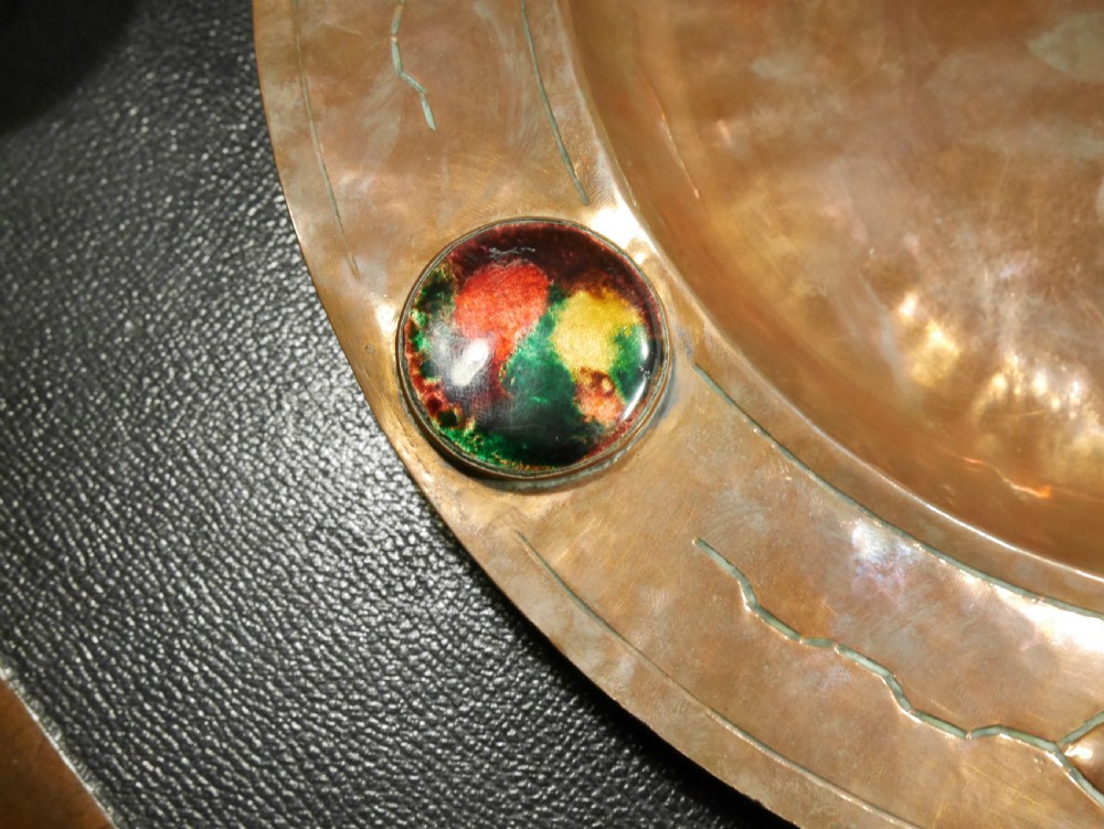 A circular hand-hammered copper charger with a sunken central dish, chased border decoration, and four inset colourful enamel cabochons. View V