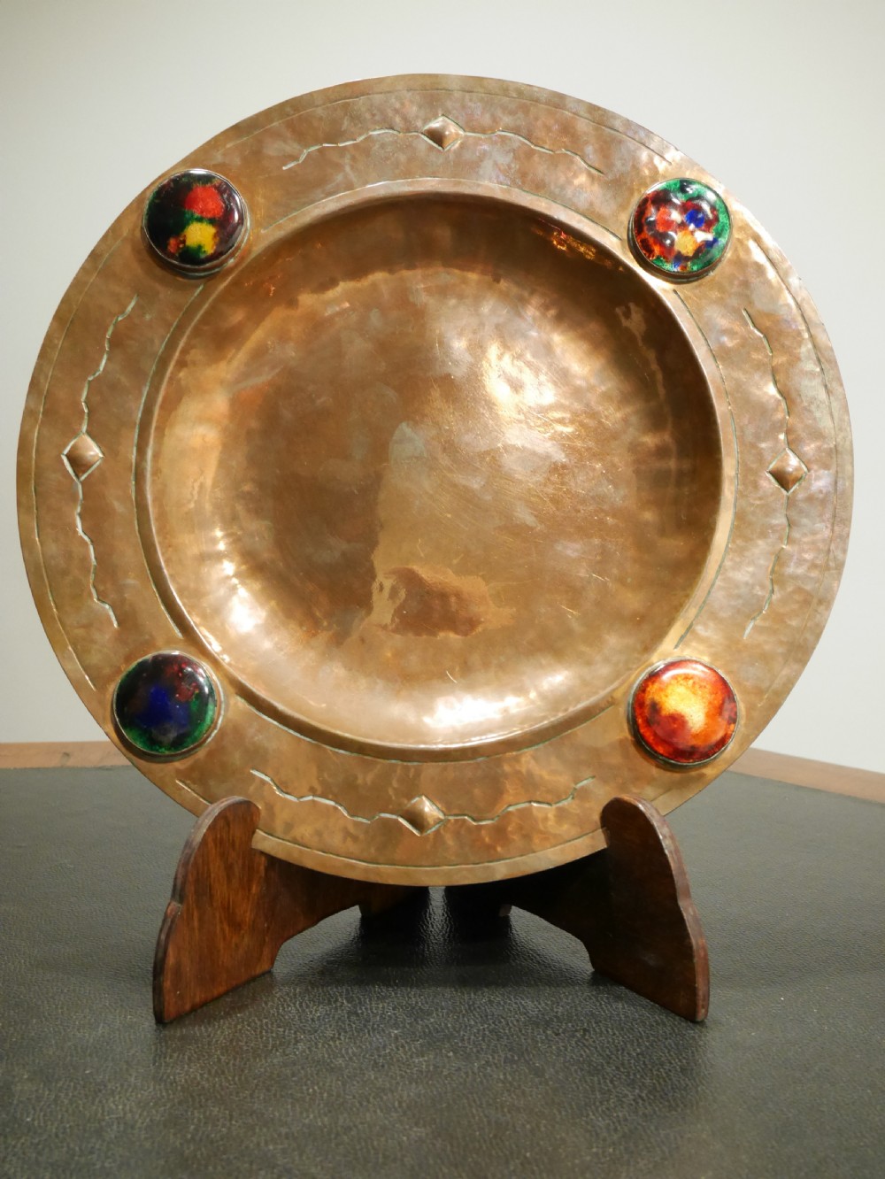 A circular hand-hammered copper charger with a sunken central dish, chased border decoration, and four inset colourful enamel cabochons. View VI