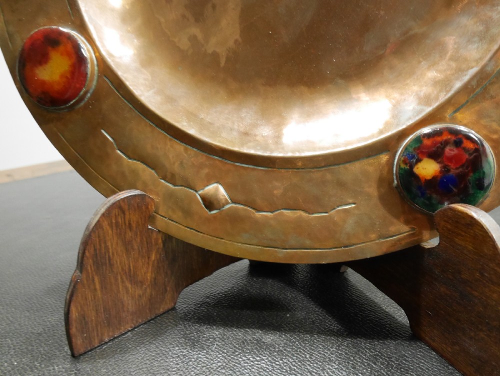 A circular hand-hammered copper charger with a sunken central dish, chased border decoration, and four inset colourful enamel cabochons. View VII