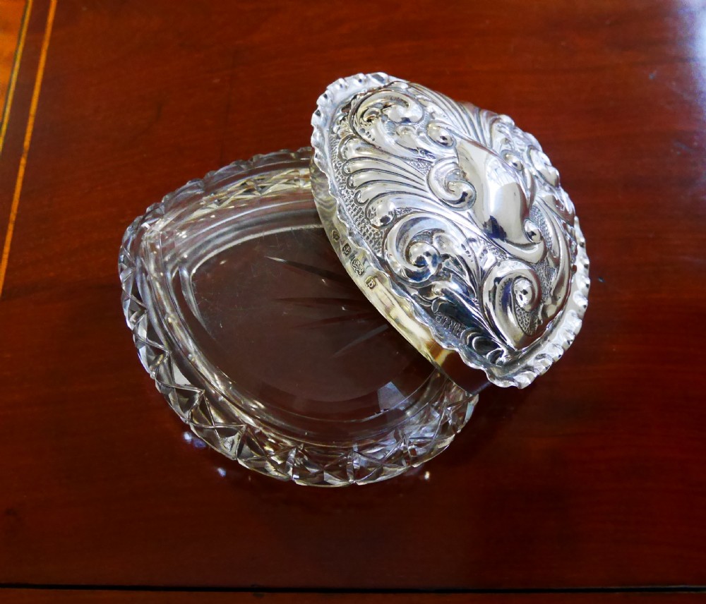 A heart-shaped cut glass trinket box featuring an embossed sterling silver lid decorated with scroll motifs. View V