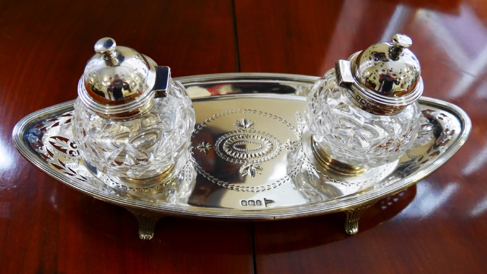 A solid silver boat-shaped tray with pierced scrollwork edges and central engraving, holding two faceted cut-glass inkwells with silver caps.