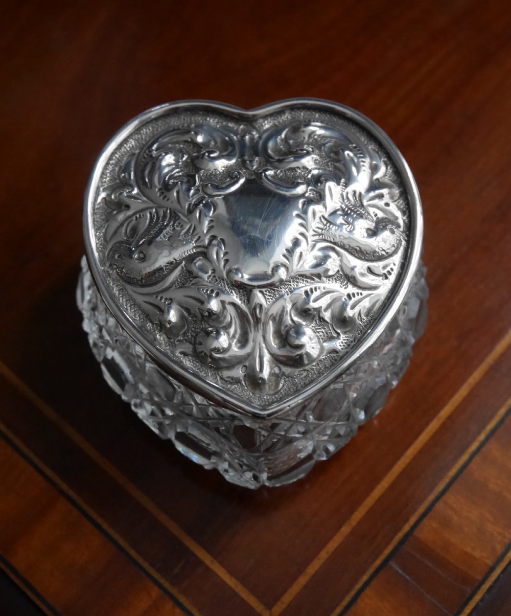 A heart-shaped cut glass trinket box with an intricately embossed silver lid featuring foliage and scrollwork motifs.