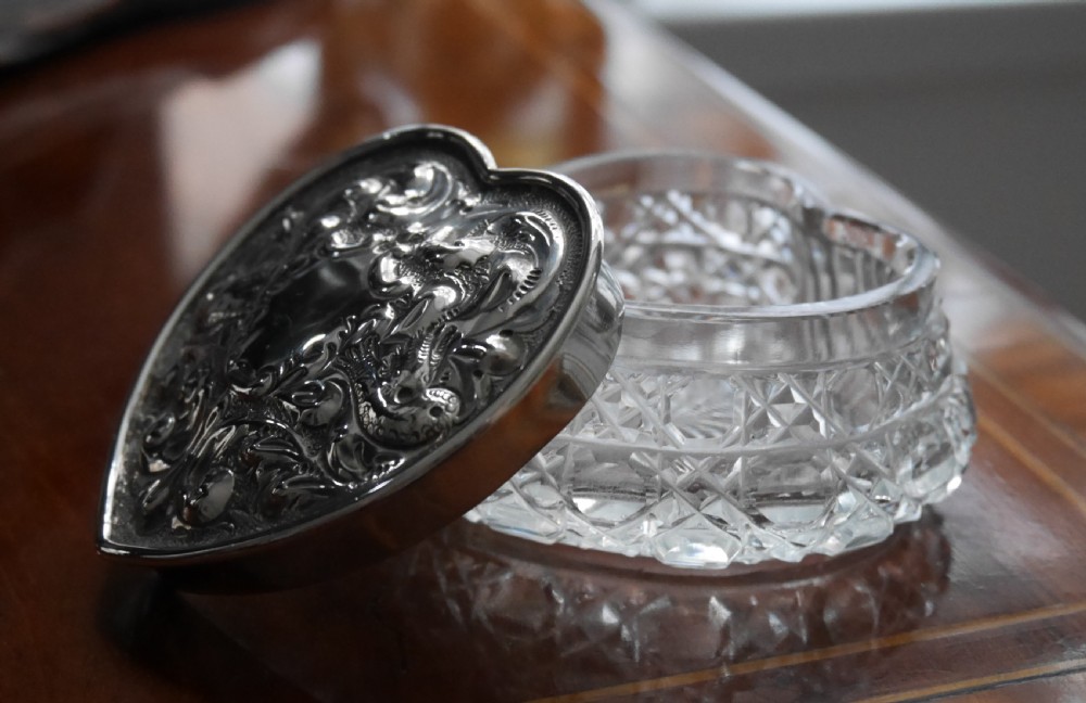 A heart-shaped cut glass trinket box with an intricately embossed silver lid featuring foliage and scrollwork motifs. View II