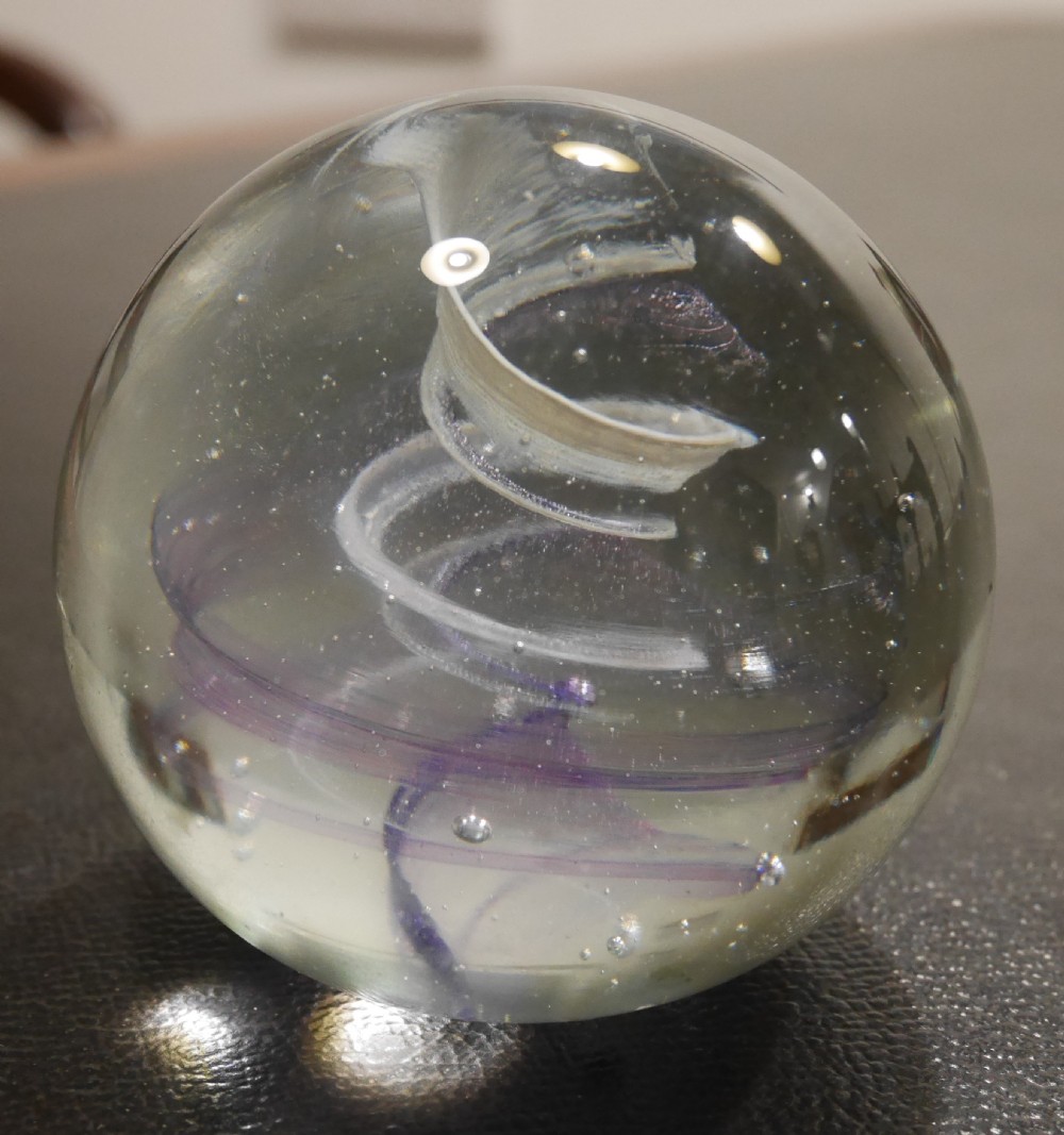 A spherical clear glass paperweight displaying an internal spiral ribbon detail in white and purple with small air bubbles.