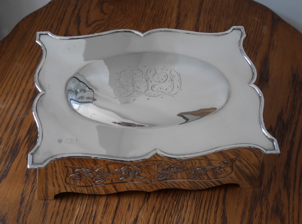 A silver rectangular jewellery casket featuring embossed floral side panels, a hinged flat lid with an engraved central monogram, and flared bracket feet. View II