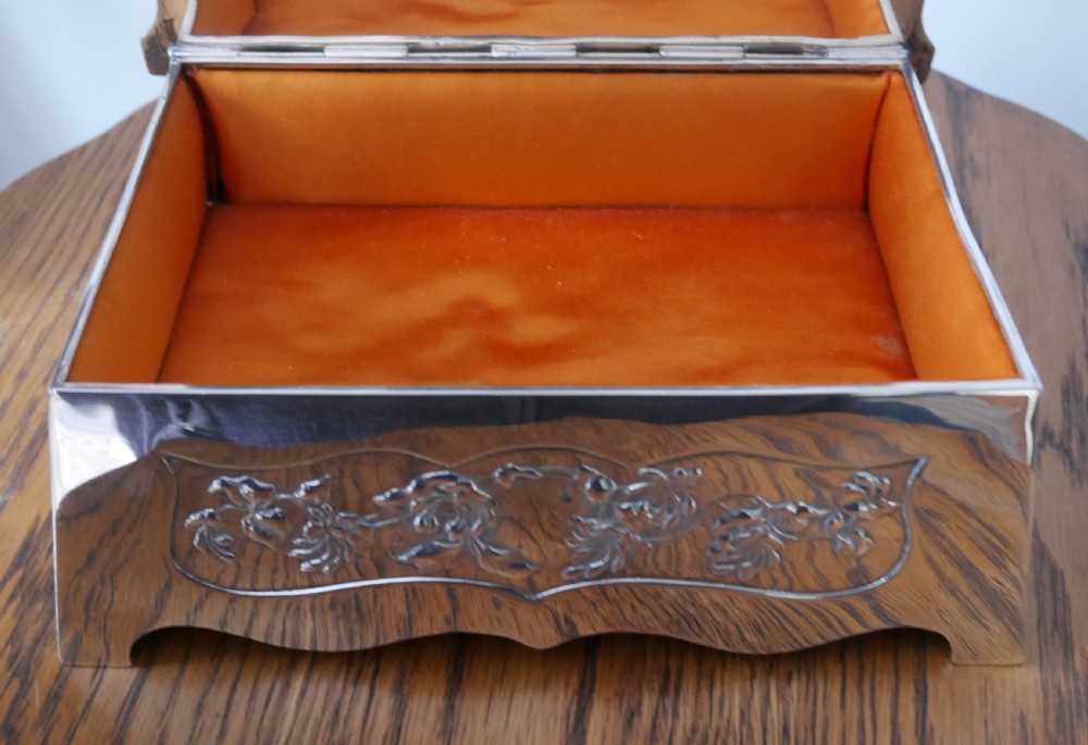 A silver rectangular jewellery casket featuring embossed floral side panels, a hinged flat lid with an engraved central monogram, and flared bracket feet. View III