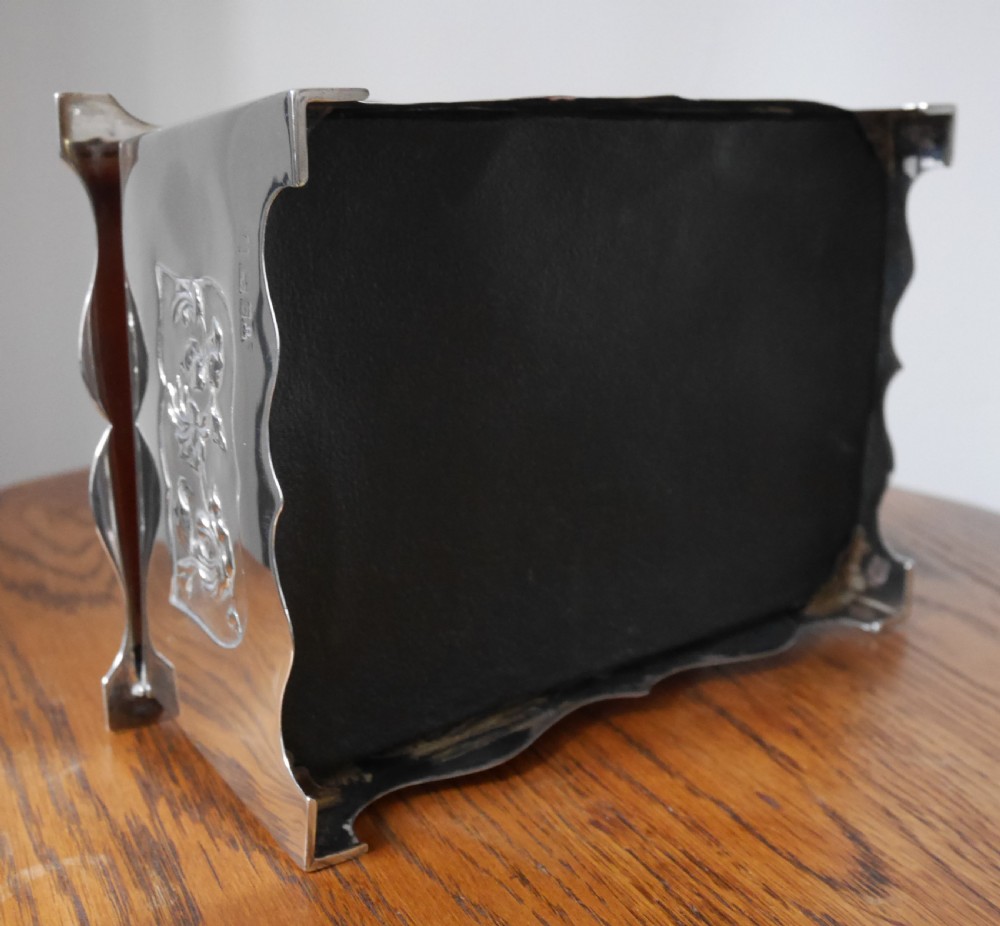 A silver rectangular jewellery casket featuring embossed floral side panels, a hinged flat lid with an engraved central monogram, and flared bracket feet. View IV