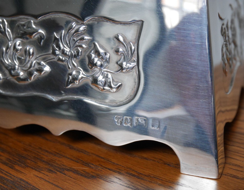 A silver rectangular jewellery casket featuring embossed floral side panels, a hinged flat lid with an engraved central monogram, and flared bracket feet. View V