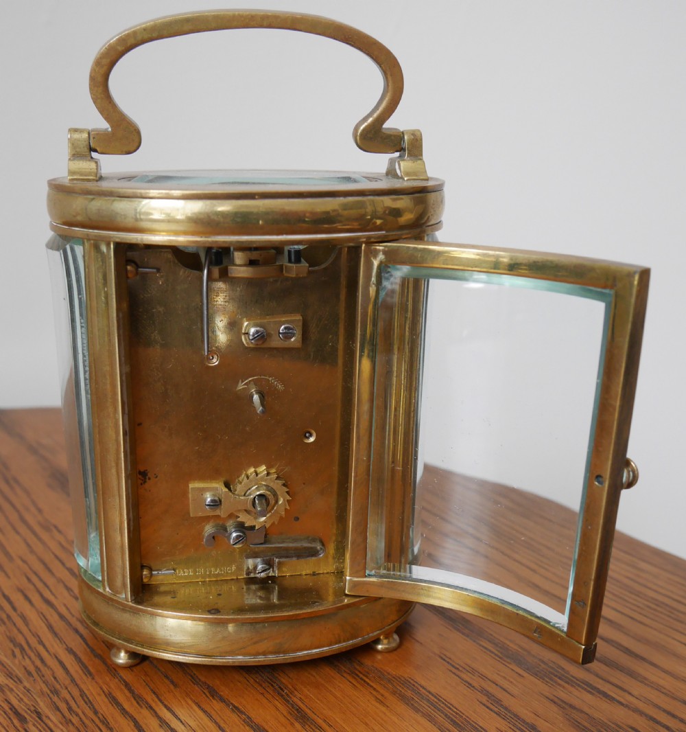 An oval brass carriage clock featuring bevelled glass side panels, a white enamel dial with black Roman numerals, and a hinged top handle. View IV