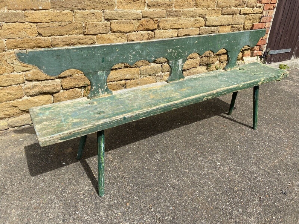 A long nineteenth-century pine bench featuring distressed green paintwork, a scalloped backrest, and splayed legs.