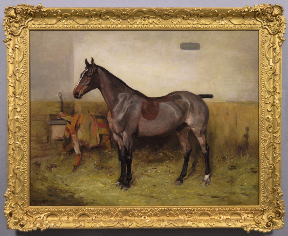 An oil painting depicting a bay hunter standing in profile inside a straw-covered stable, set within an ornate carved gilt frame.