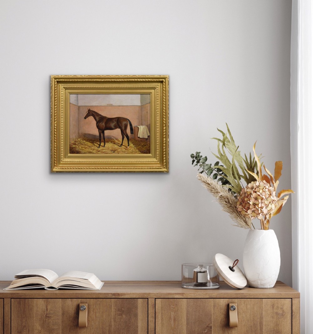 An oil painting of a brown bay racehorse standing in a straw-filled stable stall set within an ornate gold gilt frame. View IX