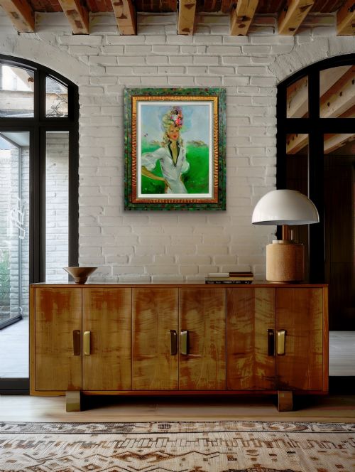 fig 9. A mid-century oil painting depicting a blonde woman in a white dress and elaborate hat standing before a green racecourse, set within a textured gilt and painted frame.