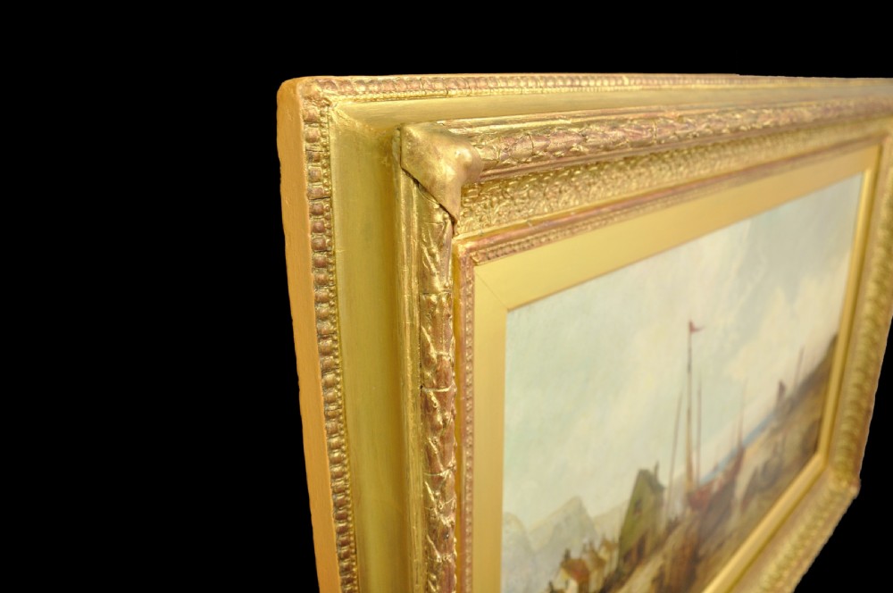 A nineteenth-century framed oil painting depicting fishing boats docked at a harbour shore beside coastal buildings, encased in a decorative gold leaf frame. View IV