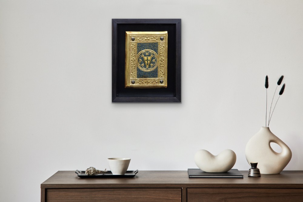 A framed rectangular gilded wooden book cover panel featuring punchwork, foliate borders, four metal corner studs, and a central coat of arms set against a blue painted background. View XX