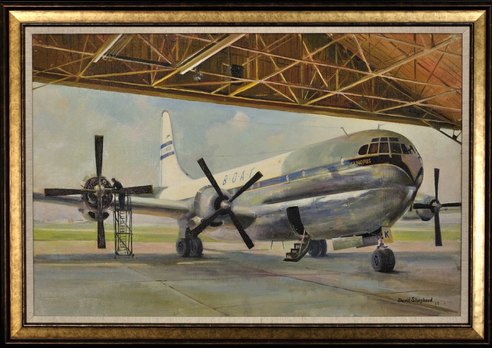 A framed oil painting depicting a BOAC Boeing Stratocruiser aircraft inside a large open hangar undergoing maintenance.