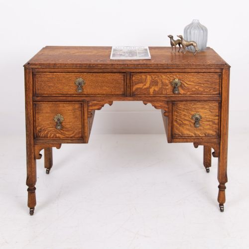 fig 1. A golden oak writing desk featuring four drawers with brass drop handles, shaped kneehole brackets, turned legs, and metal castors.