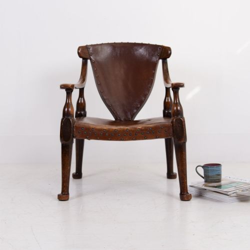 fig 1. An Arts and Crafts style dark oak armchair featuring turned front legs, wide curved armrests, and brown studded leather upholstery.