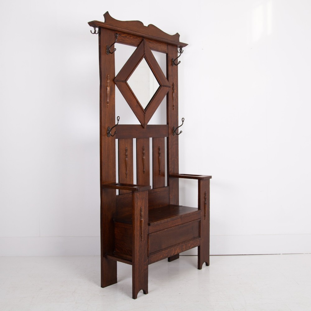 An Arts and Crafts oak hallstand featuring a diamond-shaped mirror frame, integrated brass coat hooks, turned spindles, and a seat with lift-top storage. View II