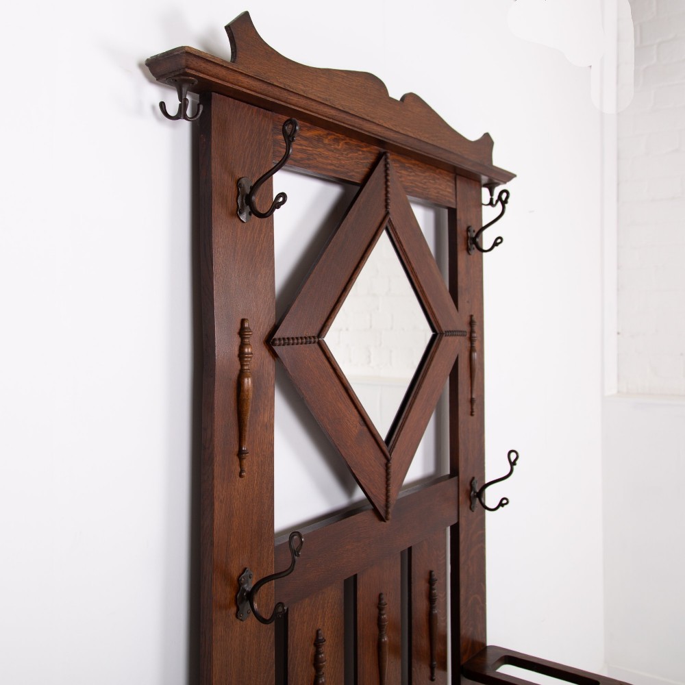 An Arts and Crafts oak hallstand featuring a diamond-shaped mirror frame, integrated brass coat hooks, turned spindles, and a seat with lift-top storage. View III