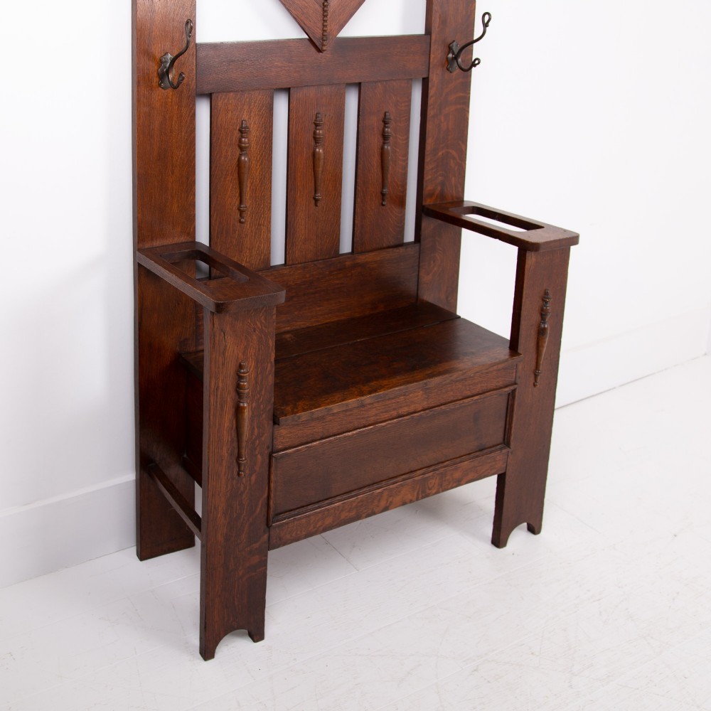 An Arts and Crafts oak hallstand featuring a diamond-shaped mirror frame, integrated brass coat hooks, turned spindles, and a seat with lift-top storage. View VI