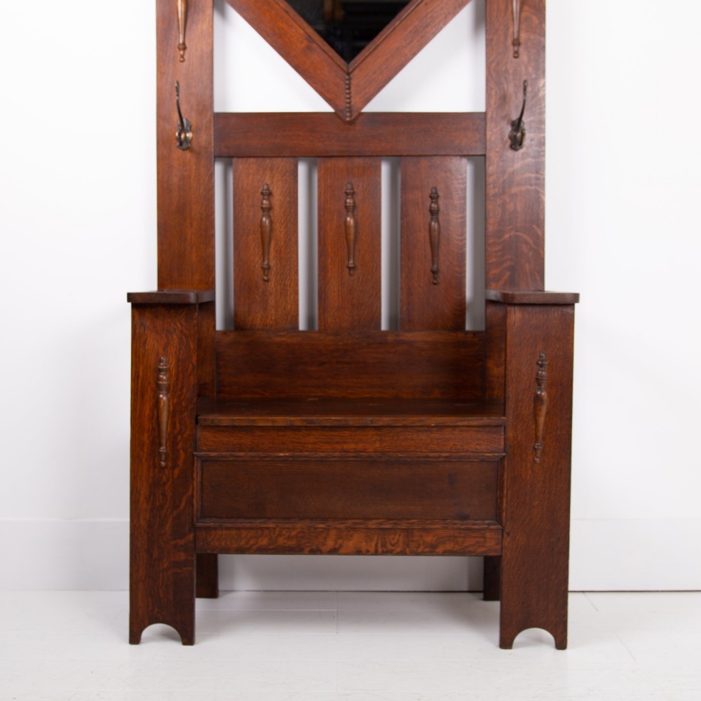An Arts and Crafts oak hallstand featuring a diamond-shaped mirror frame, integrated brass coat hooks, turned spindles, and a seat with lift-top storage. View VII