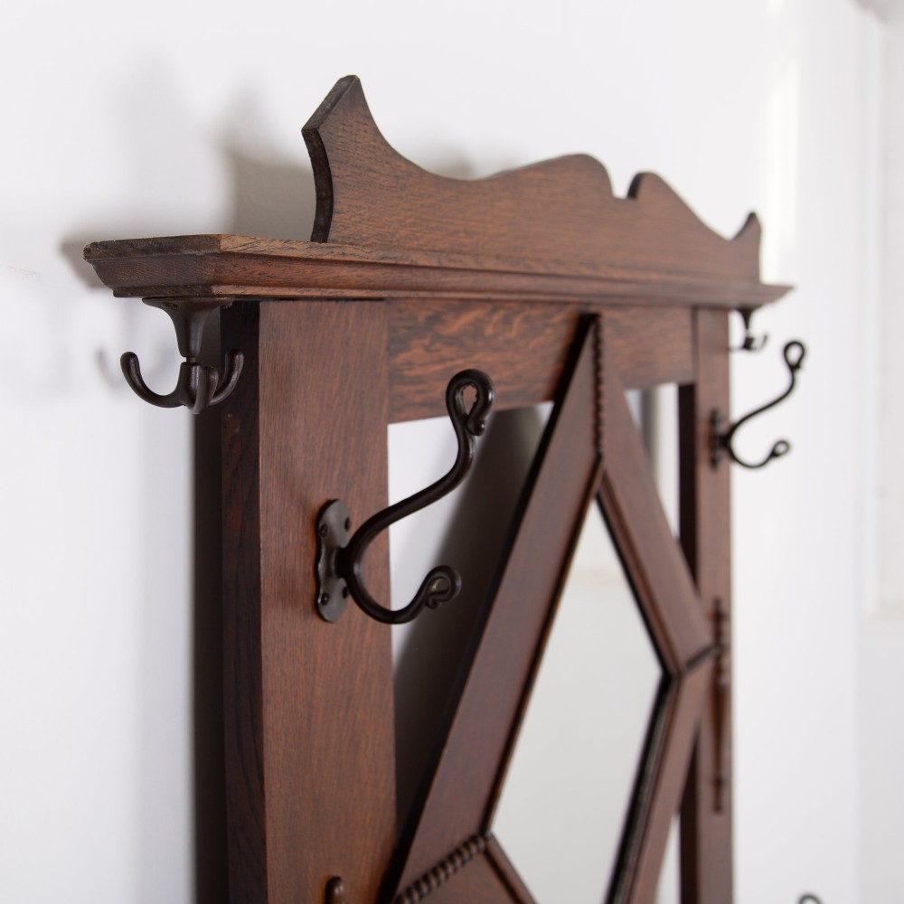 An Arts and Crafts oak hallstand featuring a diamond-shaped mirror frame, integrated brass coat hooks, turned spindles, and a seat with lift-top storage. View IX