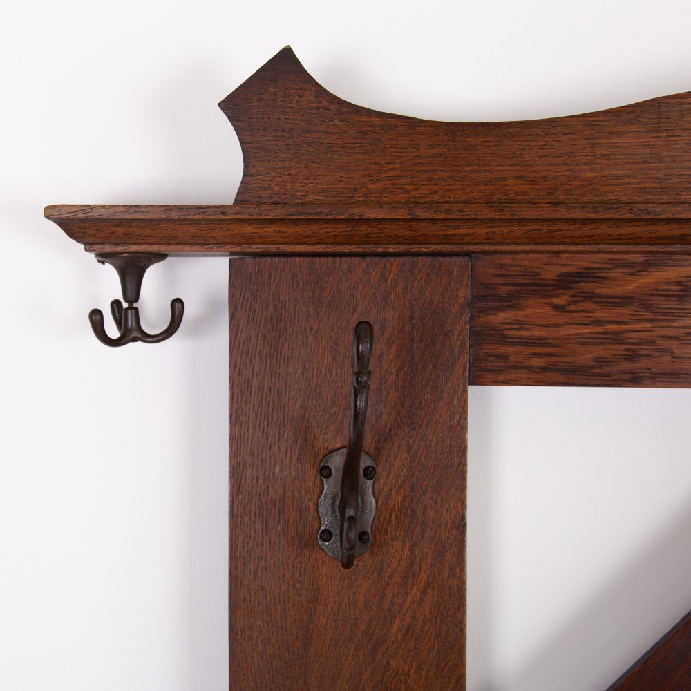 An Arts and Crafts oak hallstand featuring a diamond-shaped mirror frame, integrated brass coat hooks, turned spindles, and a seat with lift-top storage. View X