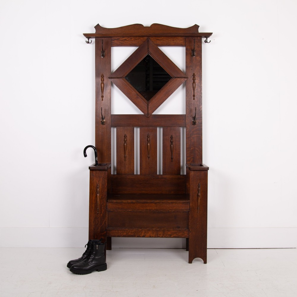 An Arts and Crafts oak hallstand featuring a diamond-shaped mirror frame, integrated brass coat hooks, turned spindles, and a seat with lift-top storage. View XII