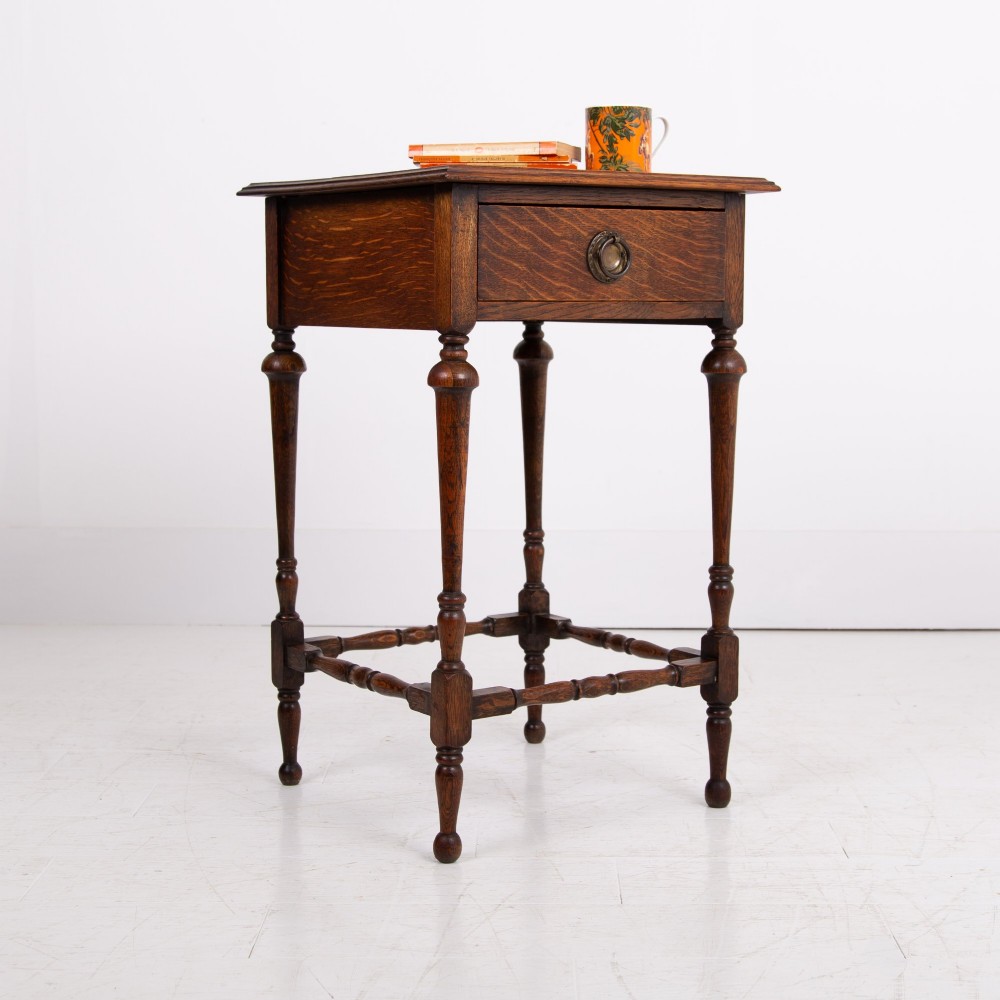 An antique dark oak side table featuring a single drawer with a brass drop ring pull, slender turned legs, and a bobbin stretcher base.