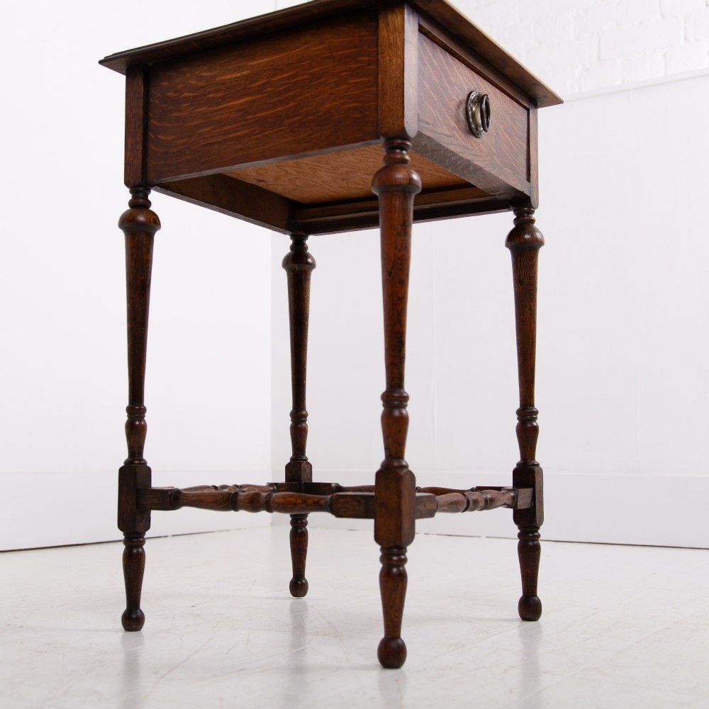 An antique dark oak side table featuring a single drawer with a brass drop ring pull, slender turned legs, and a bobbin stretcher base. View X