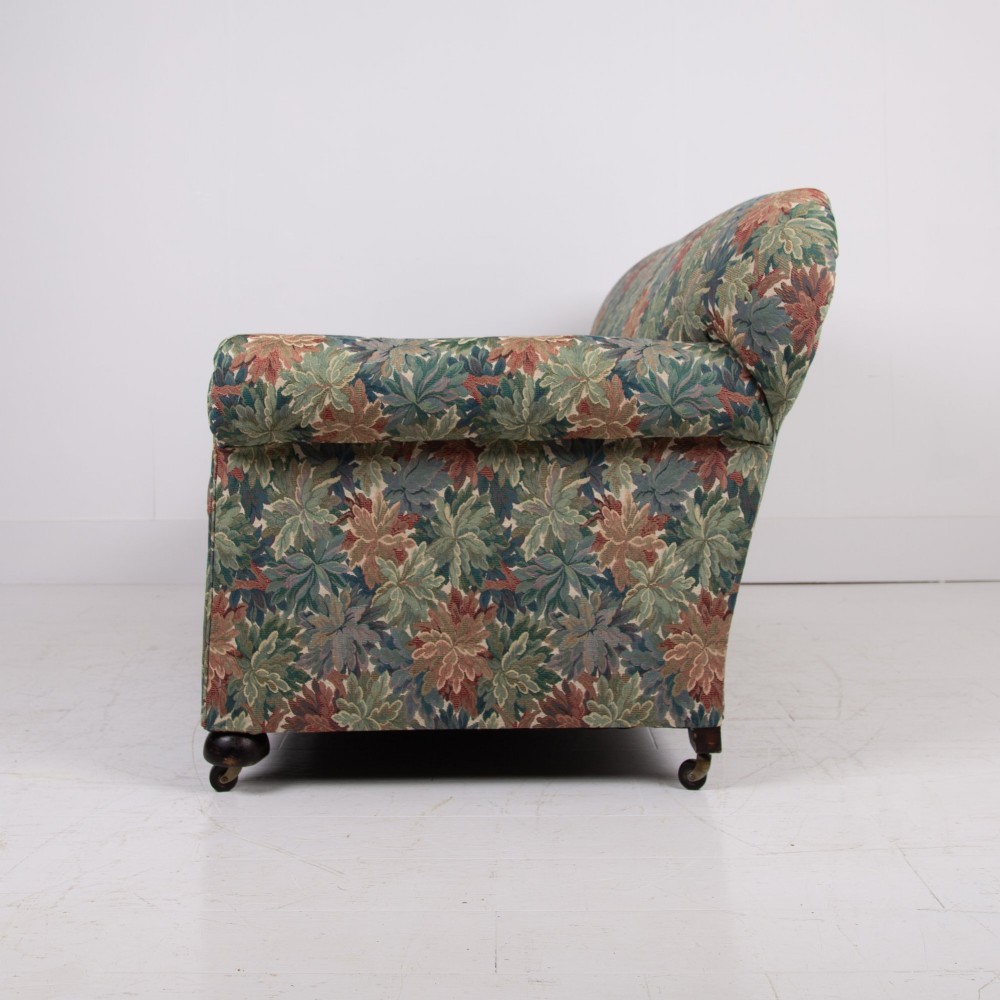An Edwardian two-seater sofa upholstered in a multi-coloured leaf tapestry fabric with rolled arms and turned timber legs on casters. View II