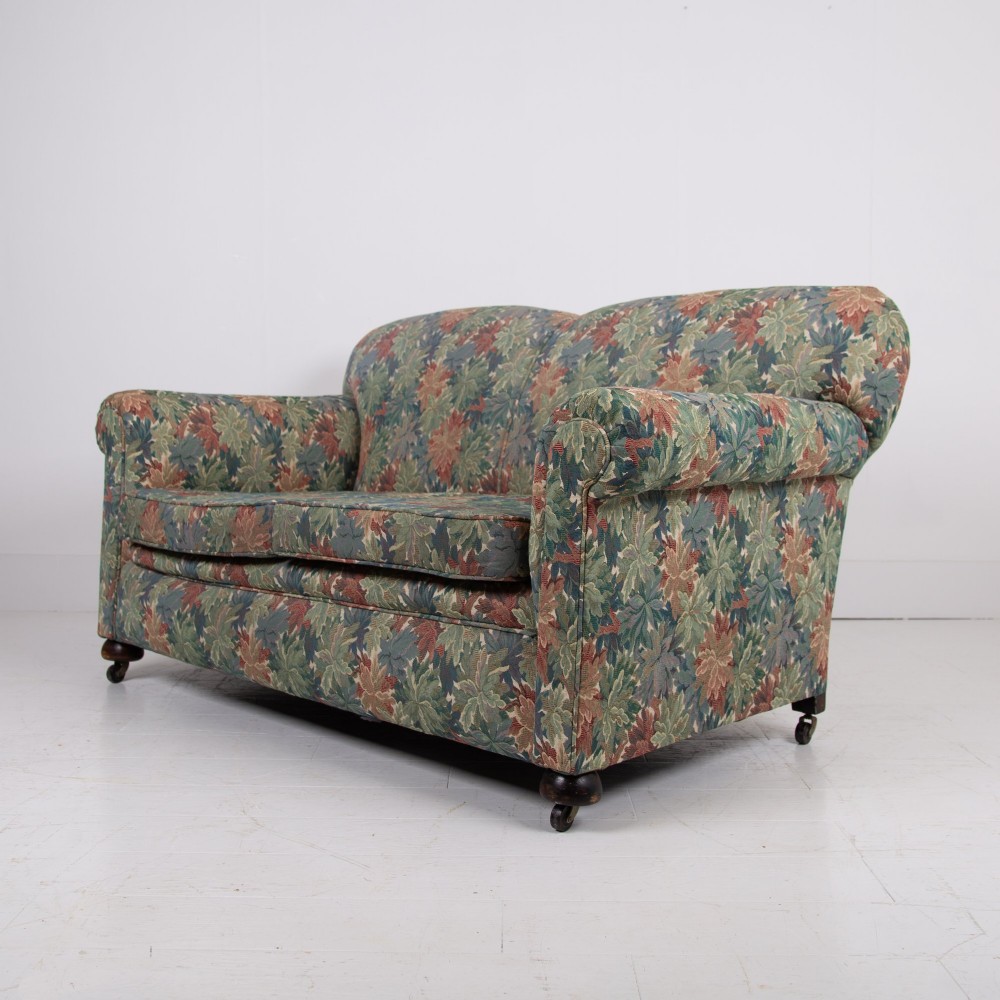 An Edwardian two-seater sofa upholstered in a multi-coloured leaf tapestry fabric with rolled arms and turned timber legs on casters. View IV