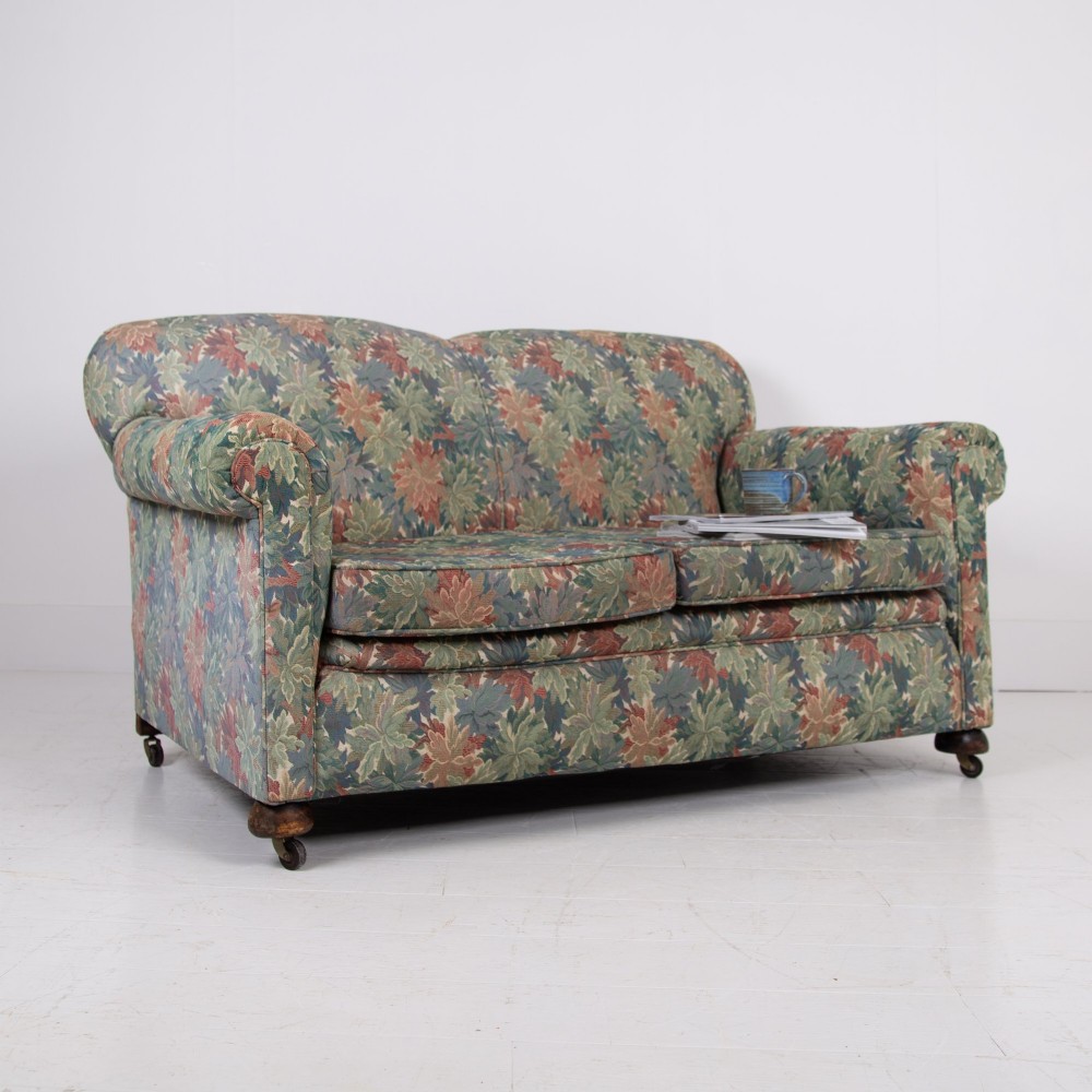 An Edwardian two-seater sofa upholstered in a multi-coloured leaf tapestry fabric with rolled arms and turned timber legs on casters. View X