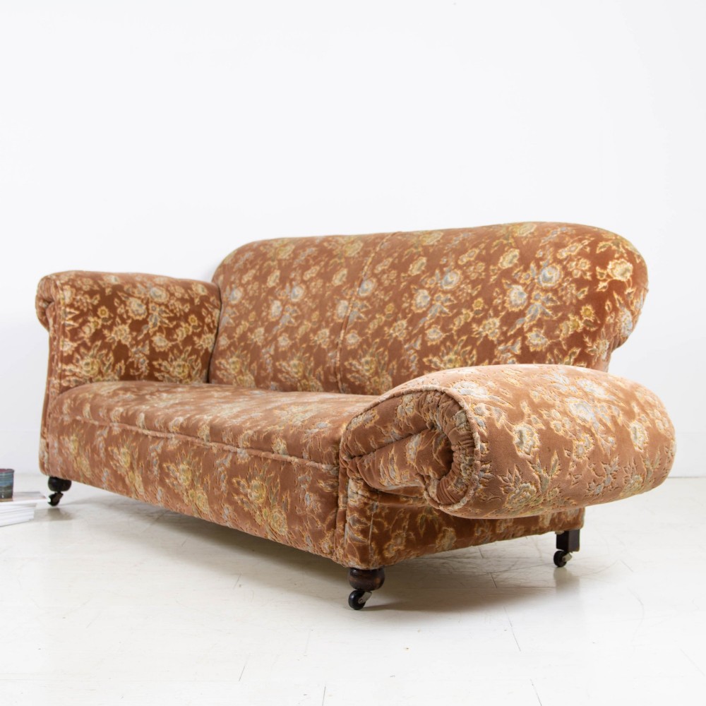 A late 1920s three-seater sofa upholstered in brown floral velvet, featuring a drop-arm end on turned wooden legs with castors.