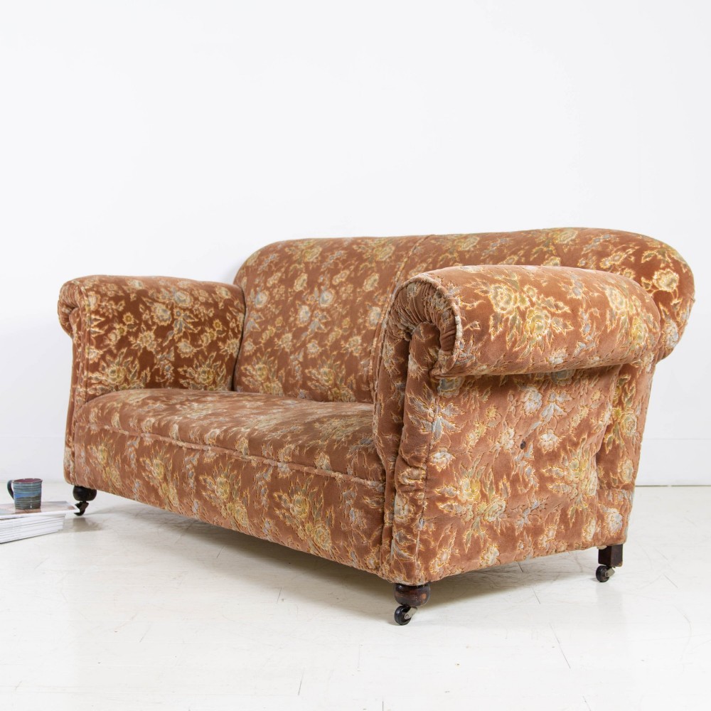 A late 1920s three-seater sofa upholstered in brown floral velvet, featuring a drop-arm end on turned wooden legs with castors. View V