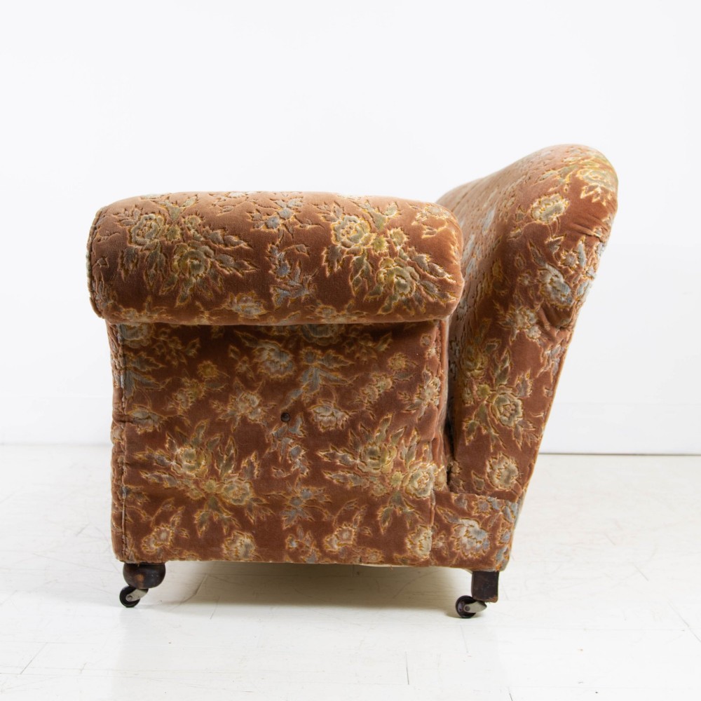 A late 1920s three-seater sofa upholstered in brown floral velvet, featuring a drop-arm end on turned wooden legs with castors. View VI