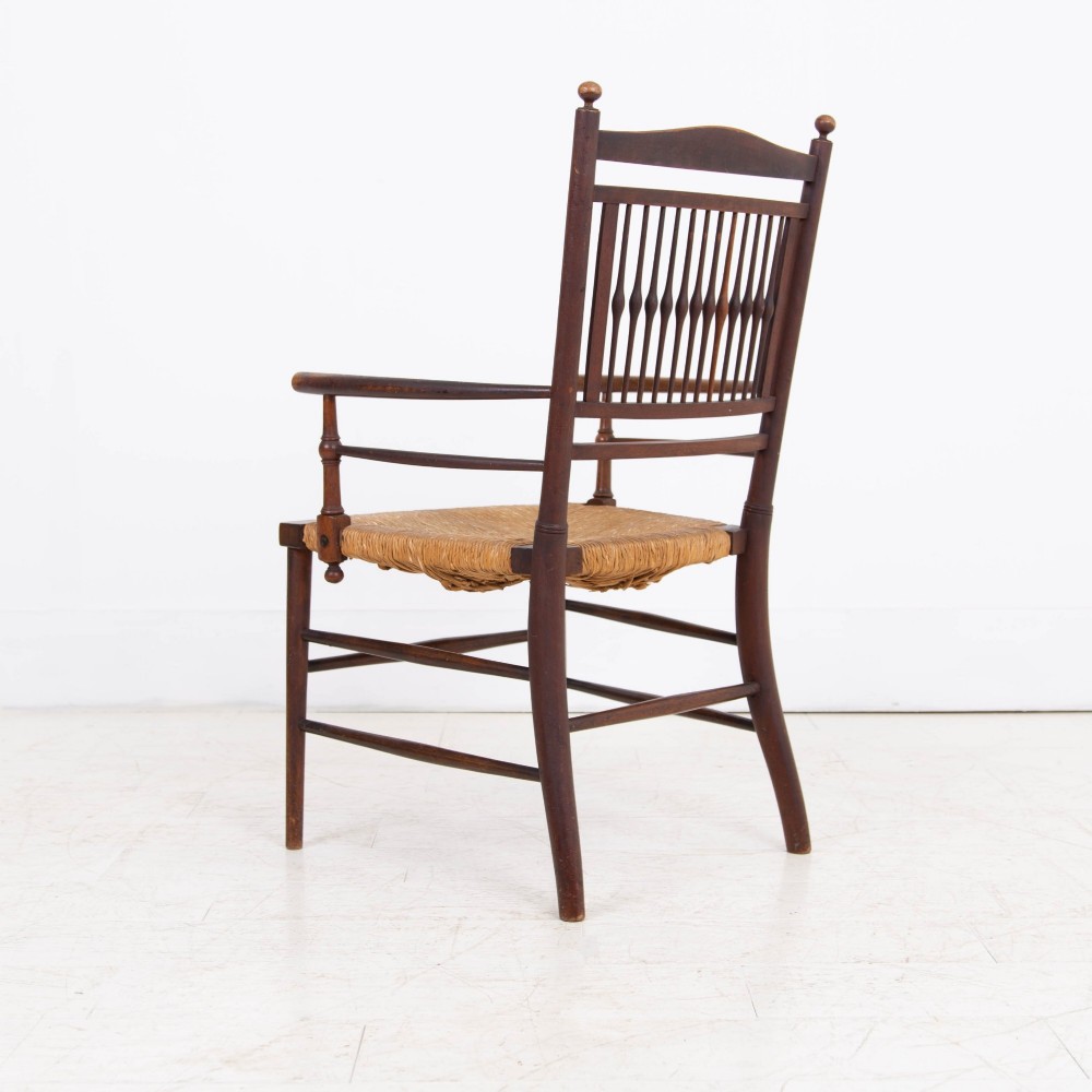 An antique Arts and Crafts wooden armchair featuring turned spindle backrest slats, sloped armrests, turned finials, and a hand-woven natural rush seat. View III