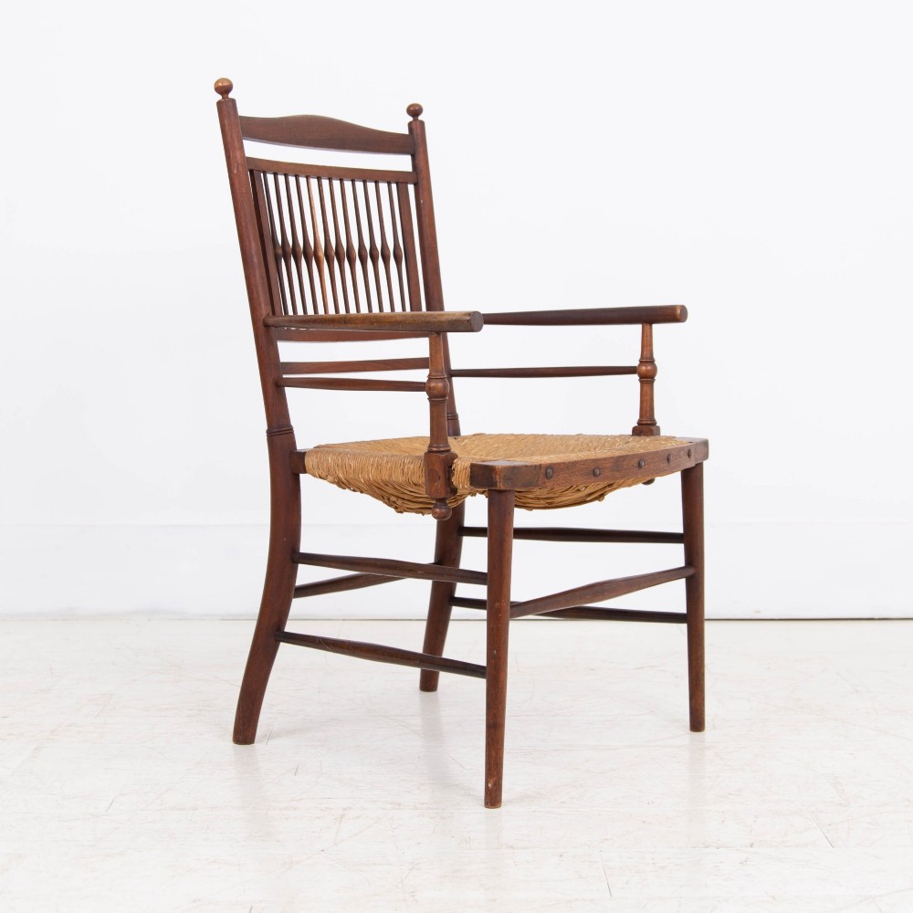 An antique Arts and Crafts wooden armchair featuring turned spindle backrest slats, sloped armrests, turned finials, and a hand-woven natural rush seat. View VII