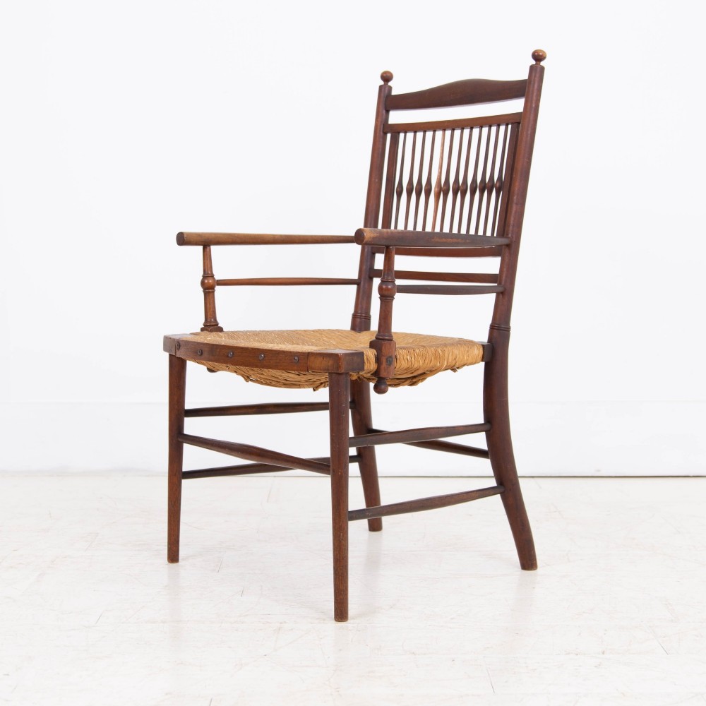 An antique Arts and Crafts wooden armchair featuring turned spindle backrest slats, sloped armrests, turned finials, and a hand-woven natural rush seat. View VIII
