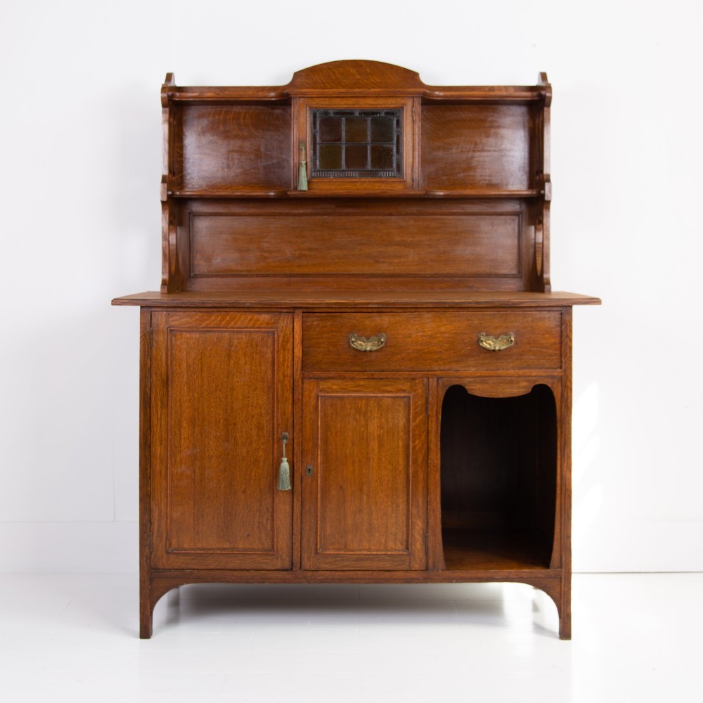 An Arts and Crafts oak cabinet dresser featuring an upper shelf unit with a central leaded glass door above a base section with a single drawer, cupboard doors, and an open side alcove.