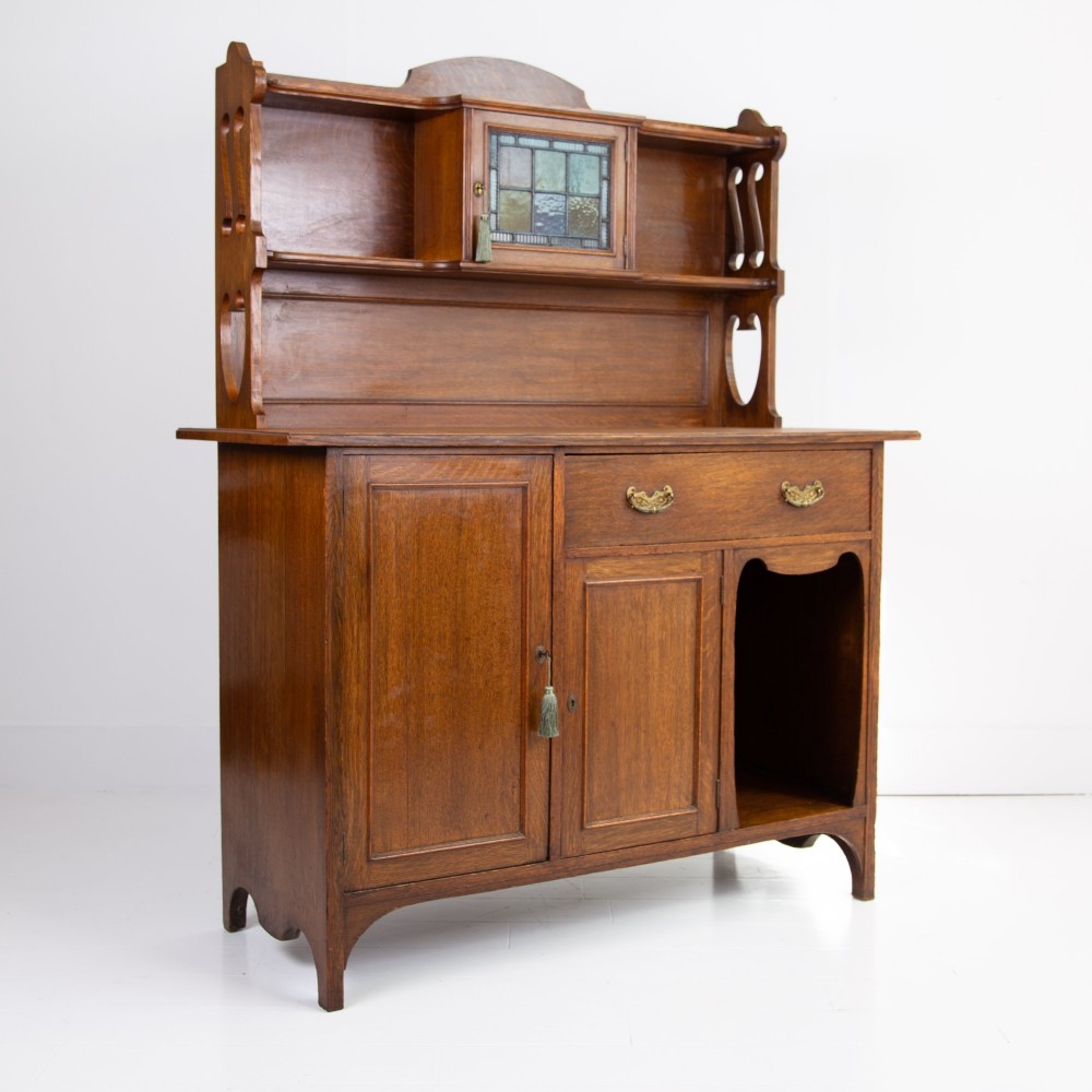 An Arts and Crafts oak cabinet dresser featuring an upper shelf unit with a central leaded glass door above a base section with a single drawer, cupboard doors, and an open side alcove. View II