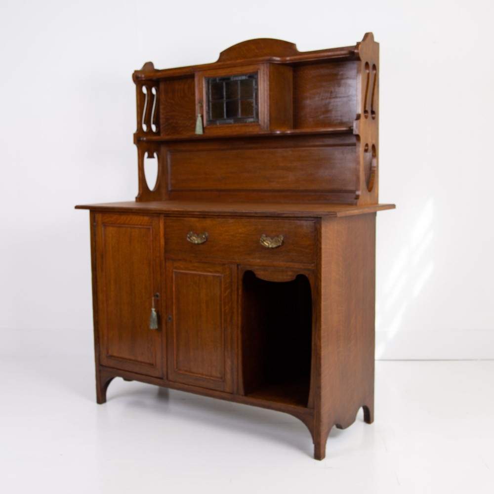 An Arts and Crafts oak cabinet dresser featuring an upper shelf unit with a central leaded glass door above a base section with a single drawer, cupboard doors, and an open side alcove. View III
