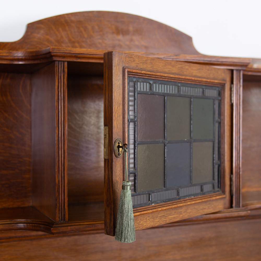 An Arts and Crafts oak cabinet dresser featuring an upper shelf unit with a central leaded glass door above a base section with a single drawer, cupboard doors, and an open side alcove. View V