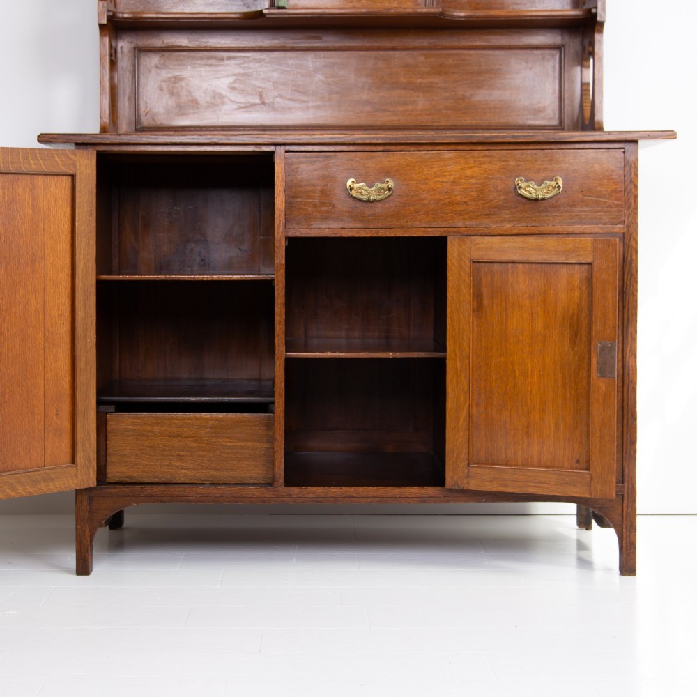 An Arts and Crafts oak cabinet dresser featuring an upper shelf unit with a central leaded glass door above a base section with a single drawer, cupboard doors, and an open side alcove. View VI